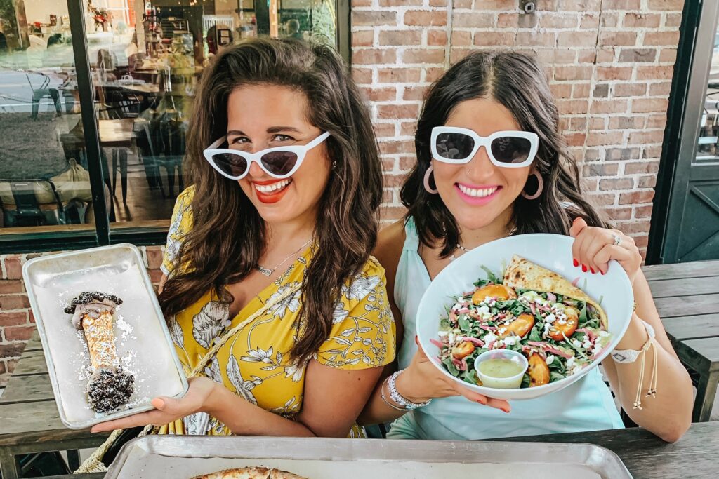 Dania and Aya in sunglasses holding a cannoli tray and a salad over two pizzas